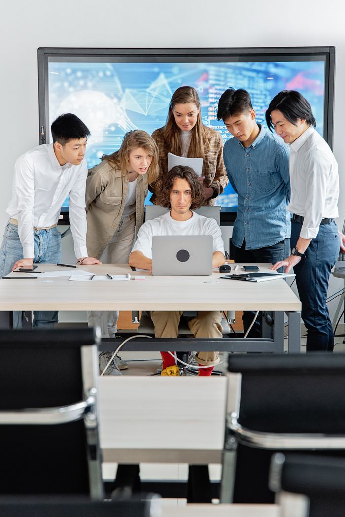 Home A diverse group of professionals engaged in teamwork around a laptop in a contemporary office setting.